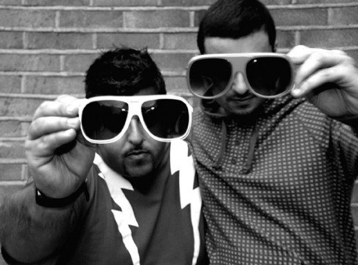 Two men holding up sunglasses in front of a brick wall.